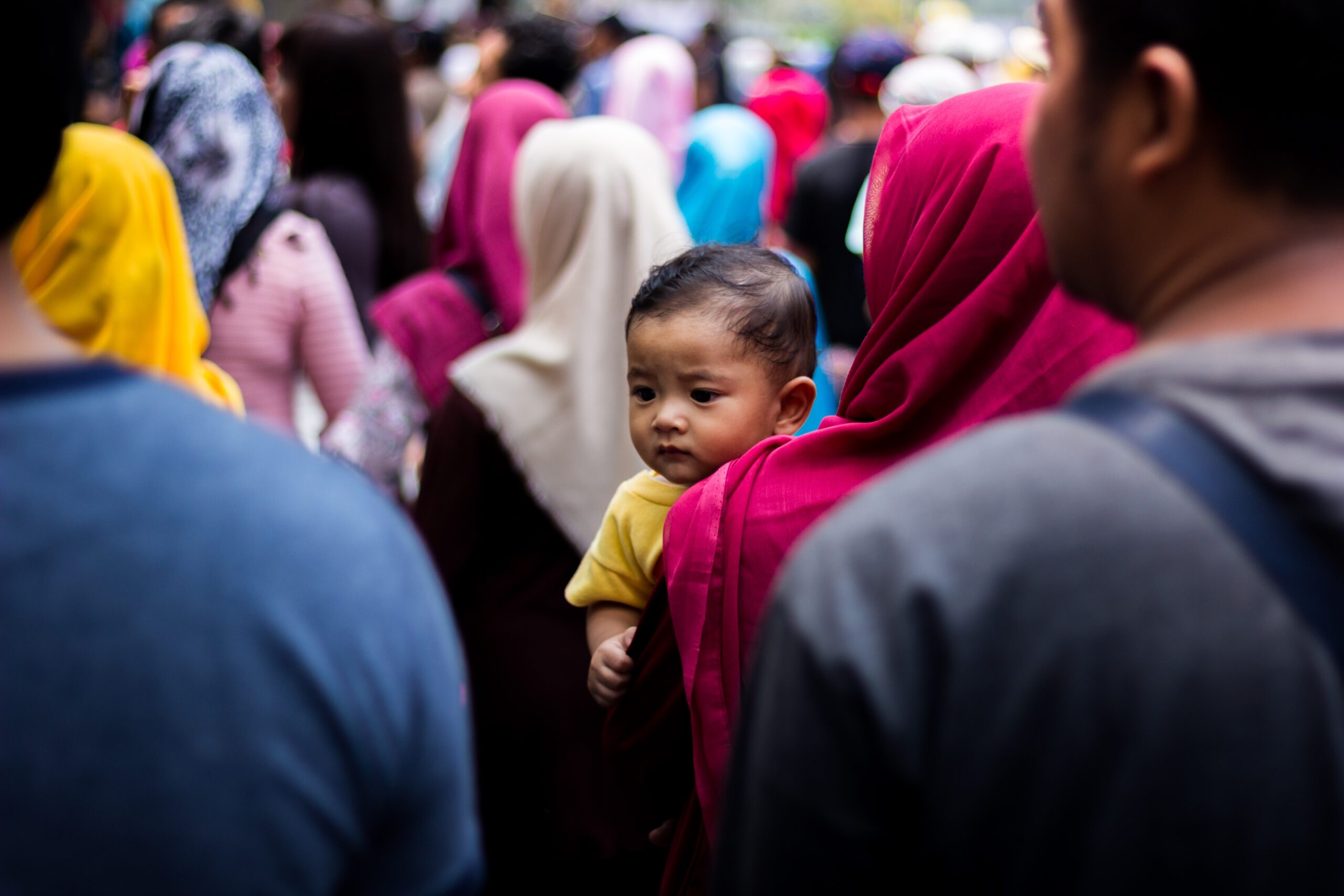 A baby wearing a yellow t-shirt on a woman’s arm in the crowd