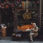 Man in Brown Jacket Sitting on Chair in front of fruit stand 